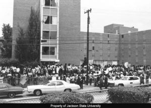 Black and white photo showing a large group of students standing outside of a multi-story concrete and brick building, a women's dormitory on campus. There are several cars parked along the street in the foreground.