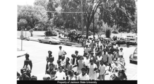 Black and white photo of a large group of students marching through streets past cars and trees.