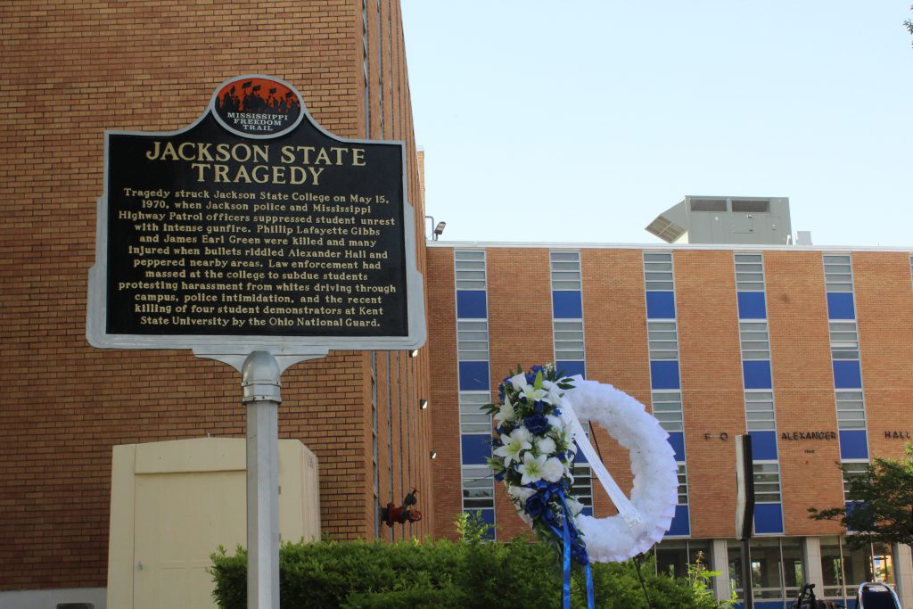 A Mississippi Freedom Trail marker sign that tells the story of the 1970 Jackson State Tragedy sits next to a floral wreath in front of a large, multistory brick building. The name of the building, Alexander Hall, is in bold letters above the doors.