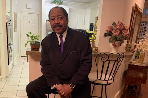 A man in a suit sits on a chair in a house, the hallway and kitchen visible beyond him.